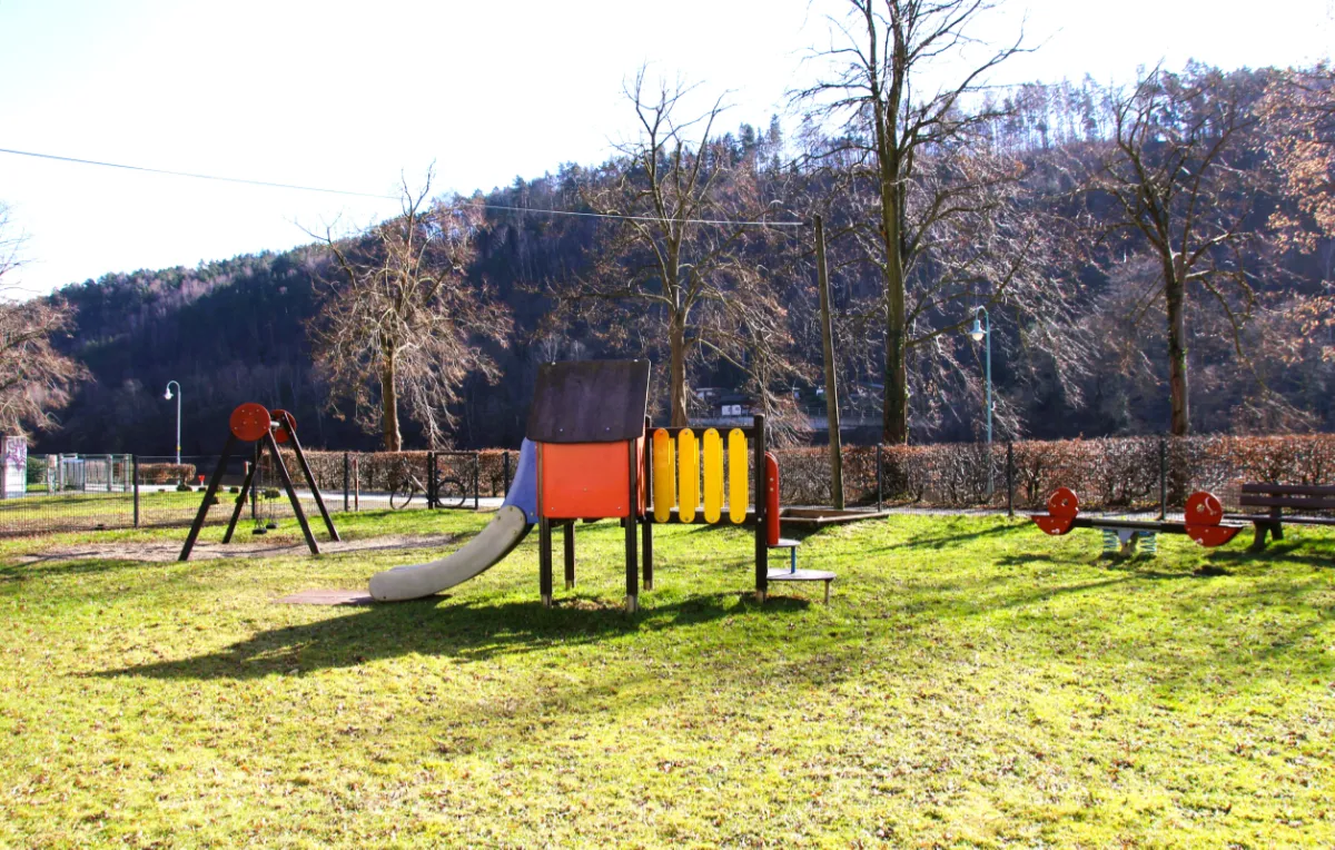 Spielplatz – Hofewiese | Foto: C. Gester Auf der Hofewiese stehen ein Klettergerüst in Rot und Gelb mit blauer Rutsche, Schaukeln, eine Wippe, ein Sandkasten und eine Bank; dahinter verläuft ein Radweg entlang einer Hecke, im Hintergrund sind kahle Bäume bei sonnigem Wetter zu sehen.