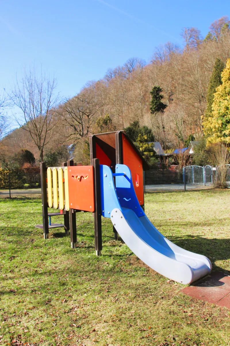 Spielplatz – Hofewiese | Foto: C. Gester Spielplatz auf der Hofewiese: Eine blaue Rutsche und andere Teile des Klettergerüsts in Rot und Gelb, auf einer Wiese, bei sonnigem Wetter; im Hintergrund kahle Bäume.